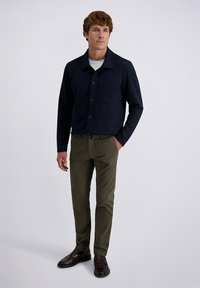 Man met kort bruin haar, gekleed in een marineblauw overhemdjack met knopen, wit ondershirt, olijfgroene broek en donkerbruine loafers, staand met één hand in zijn zak.