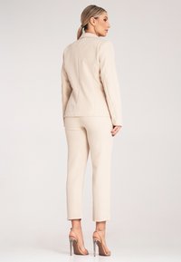 Beżowa kurtka garniturowa i wąskie spodnie wykonane z gładkiego materiału, charakteryzujące się dopasowaną sylwetką oraz eleganckim, minimalistycznym designem. Podkreślone wysokimi obcasami.