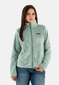 Chaqueta de forro polar verde claro con un alto cuello, patrón texturizado, cremallera frontal y una pequeña etiqueta marrón en el lado izquierdo del pecho.