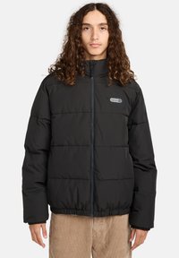 Ung person med langt krøllet hår iført en sort pufferjakke med frontlynlås og logo på brystet.