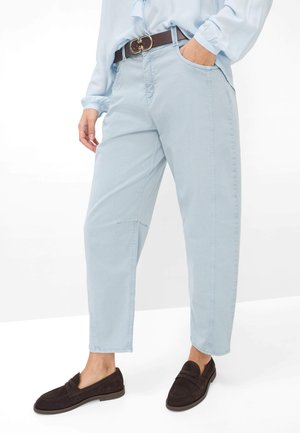 Hellblaue, zulaufende Hose, getragen mit einem braunen Gürtel, hellblauem Hemd und dunkelbraunen Slippern vor weißem Hintergrund.