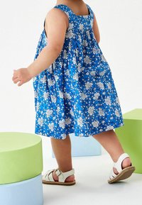 Sininen kukkamekko, jossa on valkoisia kuvioita, olkaimeton malli, gathered-back -tyyli, yhdistetty valkoisiin sandaaleihin; pastelliväristen lohkojen taustalla.