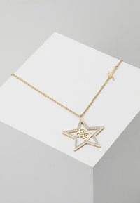 Collier chaîne en or avec un pendentif en forme d'étoile orné de petits cristaux et un logo entrelacé à l'intérieur, présenté sur une surface blanche.