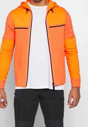 Man draagt een feloranje hoodie met ritssluiting en zwarte details, een wit shirt daaronder en zwarte cargobroek, tegen een witte achtergrond.