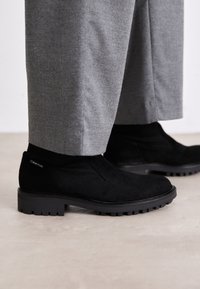 Juoda suede medžiagos auliniai batai su guma padėkliuku, turintys elastingą šoninį ketvirčius ir logotipo akcentą ant kulno. Su derančiomis pilkomis kelnėmis.
