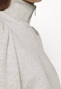 Sudadera gris de cuello alto con un cuello acanalado, que presenta un cierre de media cremallera y un tejido suave y texturizado; detalle de mangas ligeramente abullonadas.