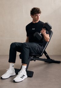 Svart trenings-T-skjorte med logo, svarte bukser, hvite joggesko med svarte detaljer, holder en sekskantet hantel mens han sitter på en vektrampe.