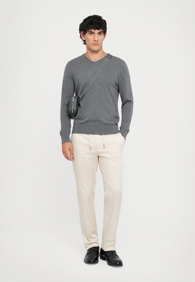 Grijze v-hals trui, crèmekleurige broek met elastische taille, zwarte instapschoenen. Model draagt een crossbody tas met ritssluiting, met een gladde textuur.