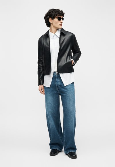 Άνδρας που φοράει μαύρο δερμάτινο μπουφάν, λευκό πουκάμισο, φαρδιά μπλε τζιν, μαύρα παπούτσια και γυαλιά ηλίου, στέκεται με το ένα χέρι στην τσέπη.