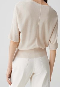 Helles beige Strickpullover mit kurzen Ärmeln, rundem Halsausschnitt und geripptem Saum. Besitzt einen subtilen Glanz und eine Mittelnaht, die entlang des Rückens verläuft.