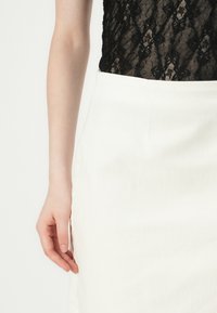 Bodysuit en dentelle noire, jupe blanche taille haute, texture lisse, design ajusté. Une main repose sur la jupe, mettant en valeur les détails de couture subtils.