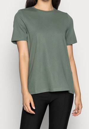 Osoba ubrana w gładki oliwkowo-zielony t-shirt z krótkim rękawem i czarne spodnie, stojąca na jasnym tle.