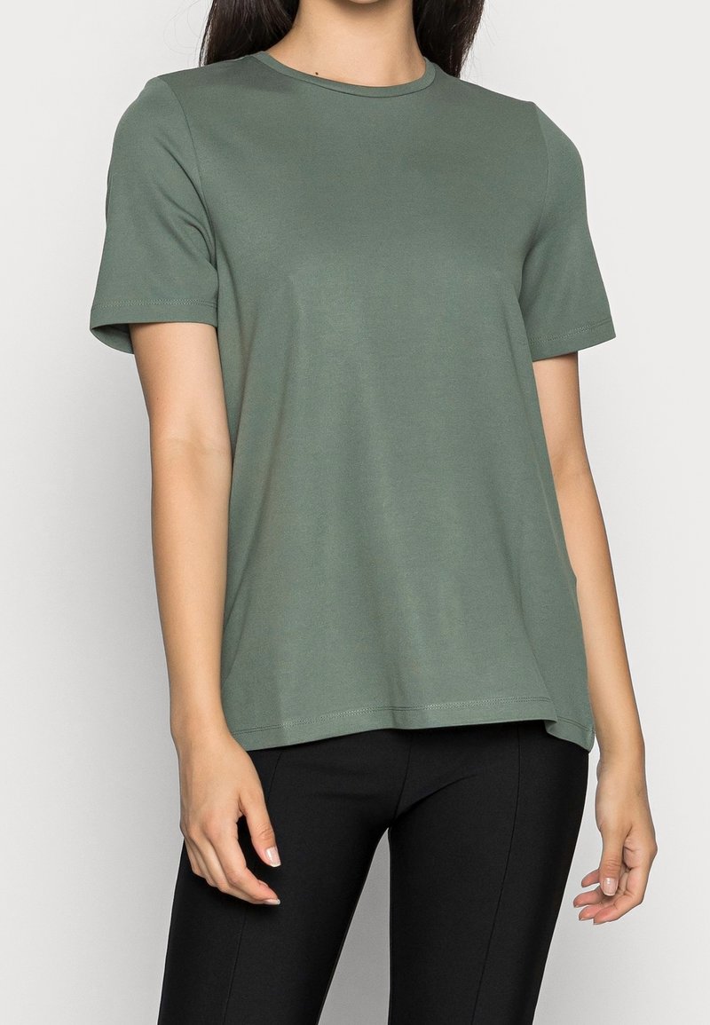 Personne portant un t-shirt vert olive uni à manches courtes et un pantalon noir, debout devant un fond clair.