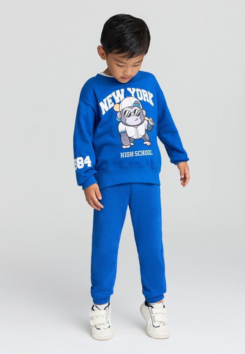 Felpa blu con il testo "NEW YORK HIGH SCHOOL" e un gorilla in stile cartone animato. Pantaloni della tuta blu abbinati. Sneakers bianche con cinturini.