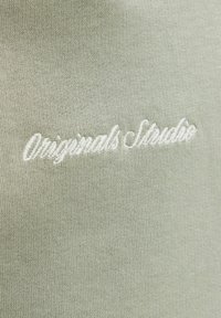 Svetlo zelena tkanina s fino teksturo, z belim vezenim besedilom "Originals Studio" v pisani pisavi.