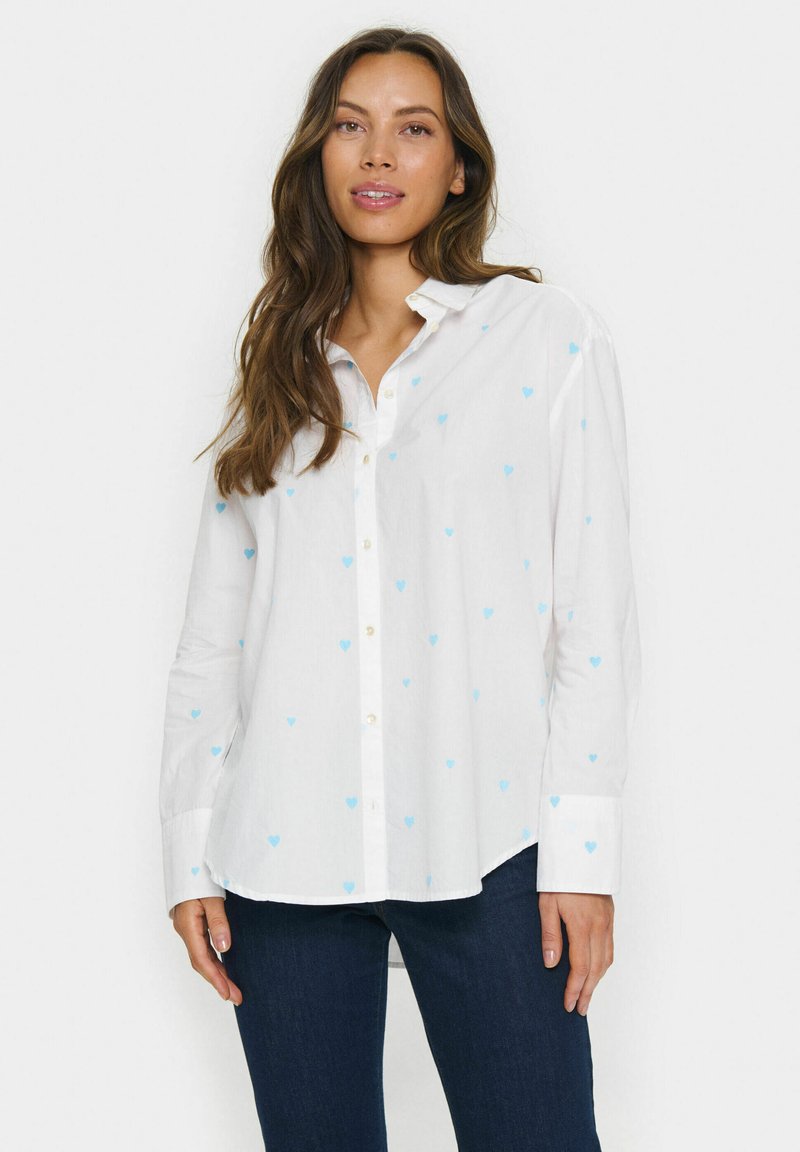 White button-up shirt featuring light blue heart patterns, long sleeves, and a curved hem. Material appears soft with a smooth texture.