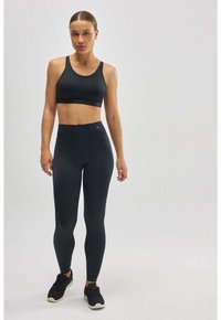 Sujetador deportivo negro con tirantes anchos y detalle de costura, combinado con leggings negros de tiro alto. La modelo lleva zapatillas deportivas negras.