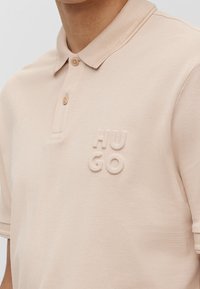 Beżowa koszulka polo z fakturowanej tkaniny, z trzema guzikami z przodu i wytłoczonym logo "HUGO" na klatce piersiowej.
