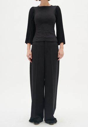 Kobieta ubrana w dopasowaną czarną bluzkę z plisowanymi długimi rękawami oraz szerokie czarne spodnie, stojąca na białym tle.