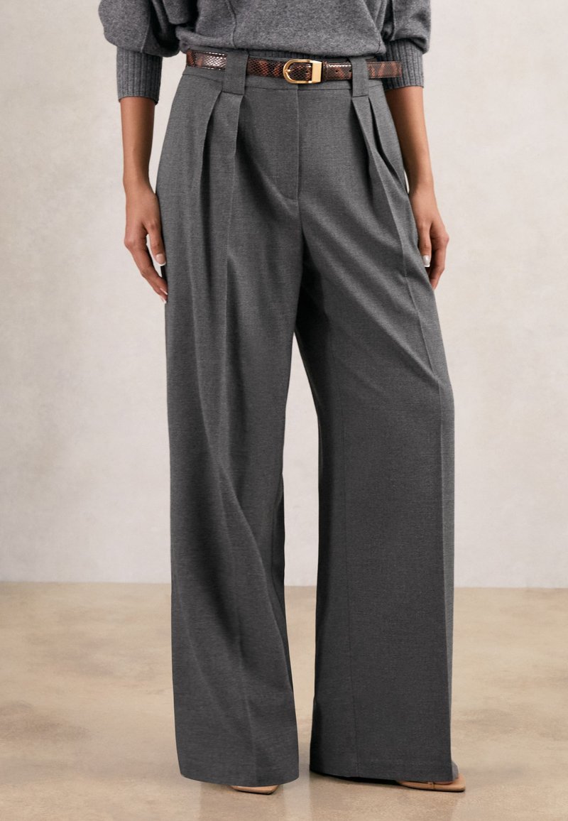 Wide-leg grey trousers with front pleats, paired with a brown snakeskin belt and beige pointed shoes.