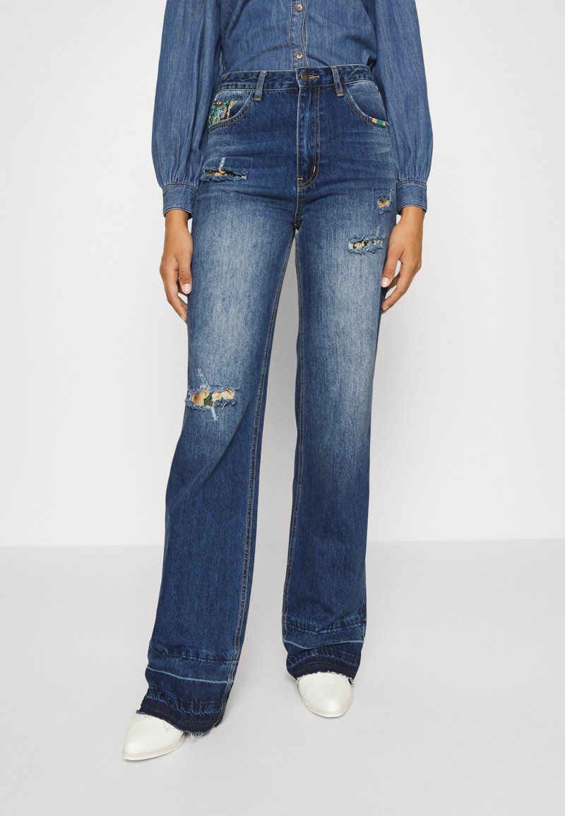 Jeans de ganga azul de perna larga com detalhes desgastados, cintura alta e bordados únicos nos bolsos. O tecido parece suave e ligeiramente desbotado.