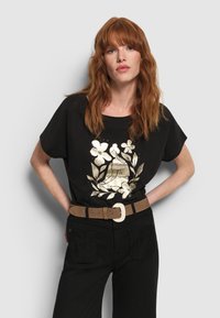Schwarzes T-Shirt aus Baumwolle mit Rundhalsausschnitt und kurzen Ärmeln, mit einem metallisch goldenen Blumenmuster in der Mitte. Brauner Gürtel mit einer großen Schnalle.