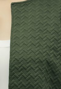 Άτομο που φοράει ένδυμα από σκούρο πράσινο υλικό με σχέδιο ζιγκ-ζαγκ, συνδυασμένο με λευκό ριμπ ύφασμα που φαίνεται στο αριστερό μέρος.