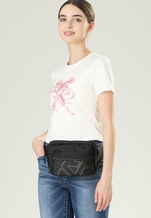 Zwarte heuptas van nylon met een gladde textuur, met een geometrisch ontwerp in wit. Draagt om de taille, gecombineerd met een wit T-shirt.