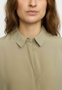 Beige shirt with a point collar, smooth texture, front button placket, and subtle stitching details around the neckline.