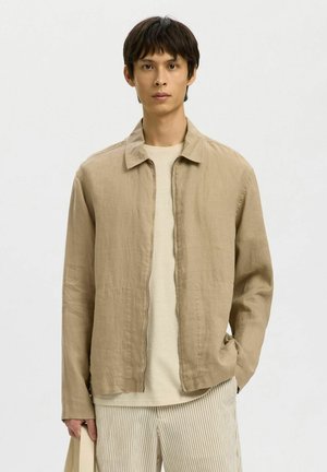 Jeune homme portant une veste en lin beige, un t-shirt crème et un pantalon à rayures claires, debout de manière détendue sur un fond uni.
