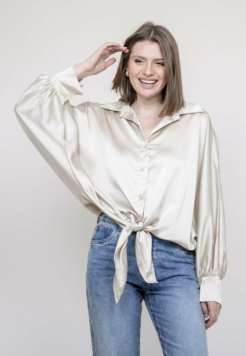 Blouse en satin crème avec un nœud à l'avant, des manches surdimensionnées et une fermeture à boutons, associée à un jean en denim bleu.