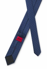 Marinblå slips i texturerad tyg med ett diamantmönster och små vita detaljer. Har en röd varumärkesetikett med "HUGO" och en ögla.