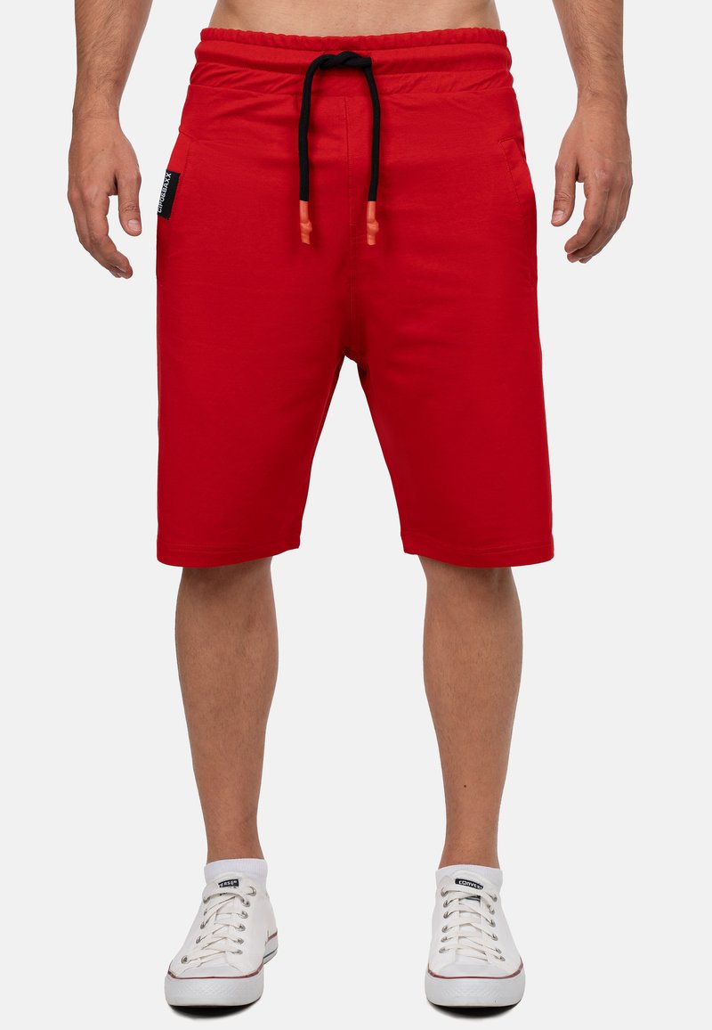 Rode katoenen shorts met een elastische tailleband en zwarte trekkoord. Voorzien van zijzakken en een zachte textuur. Draagt met een paar witte sneakers.