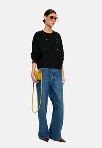 Czarny sweter dzianinowy z detalami z cekinów, niebieskie szerokie dżinsy i żółta torebka na ramię. Modelka nosi oversize'owe różowe okulary przeciwsłoneczne.