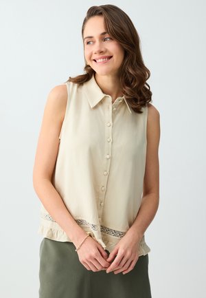 Młoda kobieta z falującymi brązowymi włosami, ubrana w beżową, bezrękawnikową bluzkę zapinaną na guziki oraz oliwkowozieloną spódnicę, uśmiechająca się i patrząca w prawo.