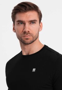 Schwarzes T-Shirt mit Rundhalsausschnitt aus Baumwolle und kurzen Ärmeln. Verfügt über ein kleines weißes "Ombre"-Logo auf der Brust. Glatte Textur, taillierter Schnitt.