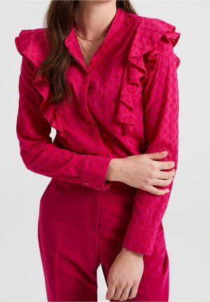 Kobieta ubrana w jasno różową bluzkę z falbanami na ramionach i fakturowanym wzorem, skompletowaną z dopasowanymi różowymi spodniami na jednolitym tle.