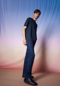 Granatowa koszulka z okrągłym dekoltem, zestawiona z niebieskimi dżinsami i czarnymi butami na sznurówki na tle gradientu od niebieskiego do różowego.