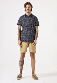 Camisa polo azul marino con un patrón de hojas, combinada con pantalones cortos caqui. Incluye mangas cortas y un cuello clásico; la persona está de pie.