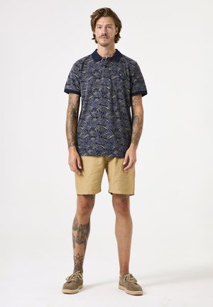 Camisa polo azul marino con un patrón de hojas, combinada con pantalones cortos caqui. Incluye mangas cortas y un cuello clásico; la persona está de pie.