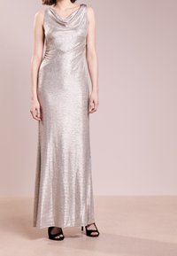 Metalická stříbrná večerní šaty až na zem s lodičkovým výstřihem a drapovaným designem, doplněné černými lodičkami s otevřenou špičkou na texturovaném povrchu.