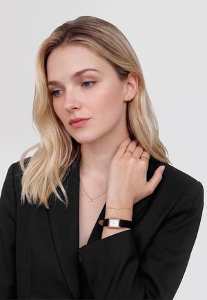 Blonde vrouw in een zwarte blazer, met gouden ringen, een fijn kettinkje, armband en een vierkante horloge met een zwarte band.