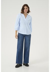 Blouse bleu clair avec un décolleté en V, motifs en forme de cœur discrets et manches bouffantes, associée à un jean en denim noir à jambes larges et des chaussures noires.