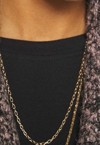 Top negro de manga larga con cuello acanalado, combinado con una cárdigan gruesa y texturizada, adornada con dos collares de cadena dorada.