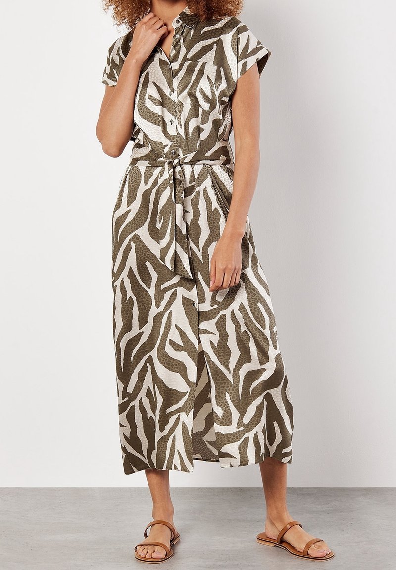 Femme portant une robe boutonnée longueur mollet avec des motifs abstraits de feuilles vertes et blanches et des sandales plates marron, debout sur un sol gris.