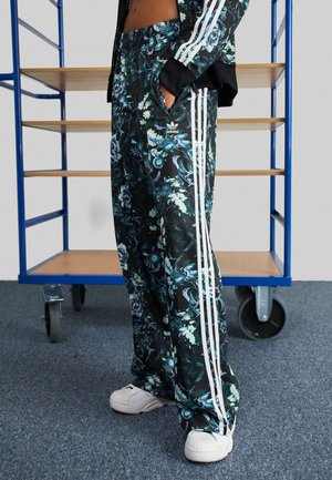 Osoba ubrana w czarne kwiatowe spodnie dresowe Adidas z białymi paskami i białe trampki, stojąca obok niebieskiego metalowego wózka na kółkach.