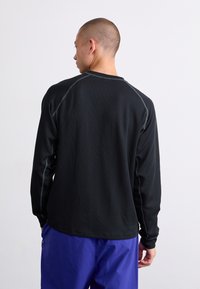 Schwarzes Langarmshirt aus einem strukturierten Stoff, mit kontrastierenden grauen Nähten und lockerer Passform, getragen mit lila Hosen.