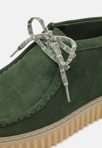 Scarpa in suede verde con superficie testurizzata, dettagli verdi coordinati e un lacci a motivi grigio e bianco. Presenta una suola chunky e traslucida.