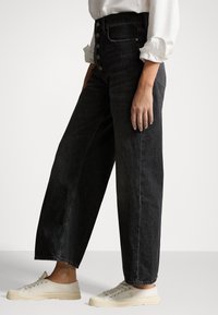 Persona vistiendo jeans de pierna ancha de tiro alto, negros, con cierre de botones, una camisa blanca de mangas arremangadas, y zapatillas de lona color crema, de pie de lado.