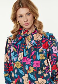 Blusa floreale con colori vivaci su uno sfondo blu scuro, colletto a fiocco, maniche arricciate e una combinazione di motivi floreali grandi e piccoli.